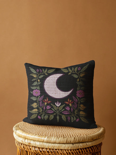 Black woven throw pillow with an embroidered crescent moon and floral pattern in soft ivory, green, purple, and gold, featuring a textured finish.